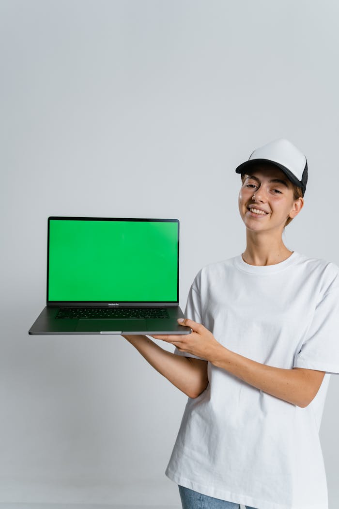Young person in white t-shirt and cap holding laptop with green screen for copy space.